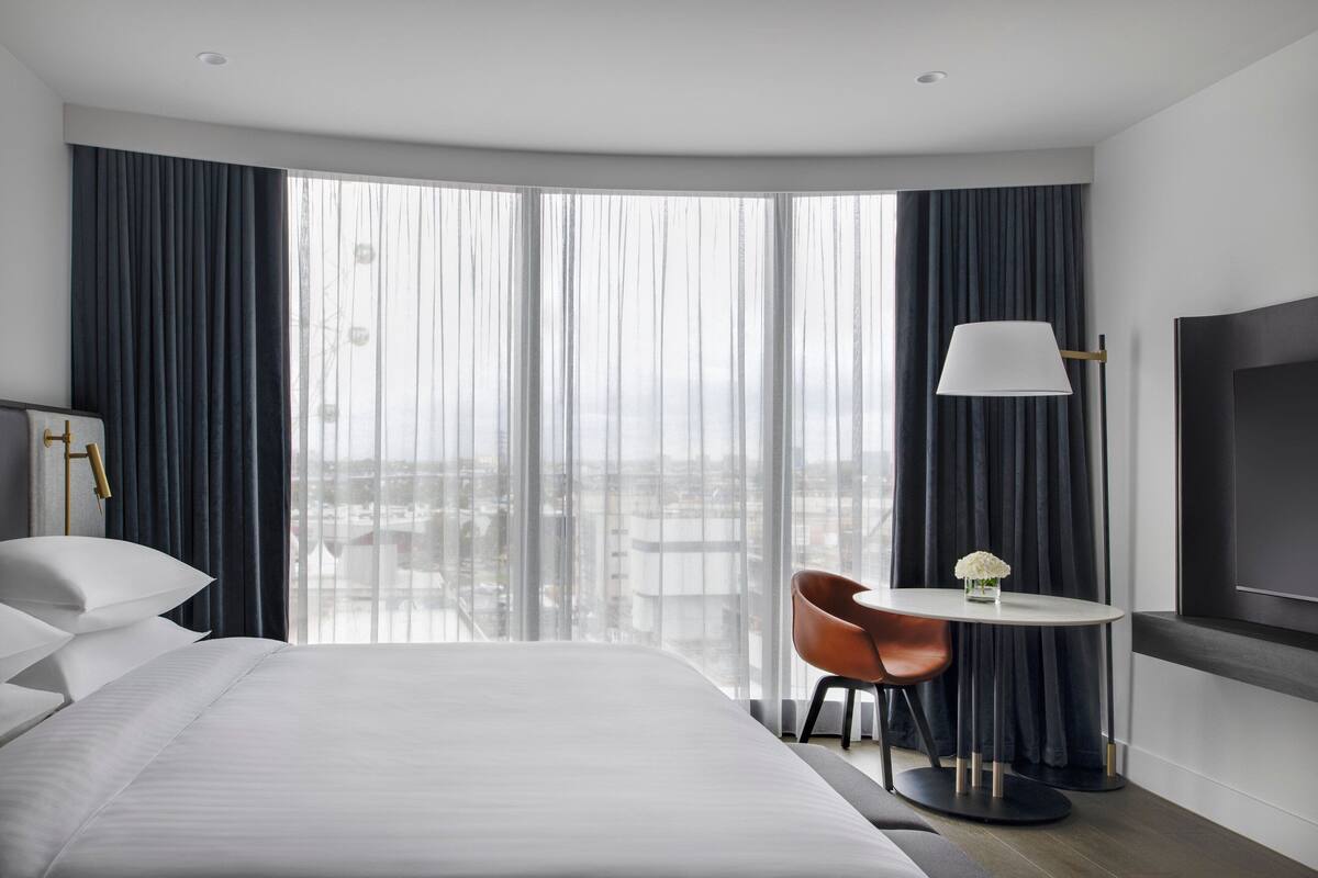 A modern hotel room with a large bed, a round table with a chair, a TV mounted on the wall, and a view of the city through large windows.
