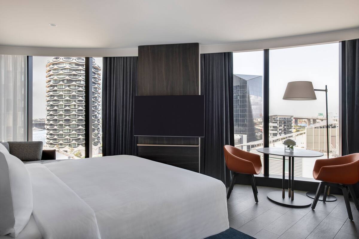 A modern hotel room with a large bed, a TV, a dining table with chairs, and a view of a cityscape through large windows.