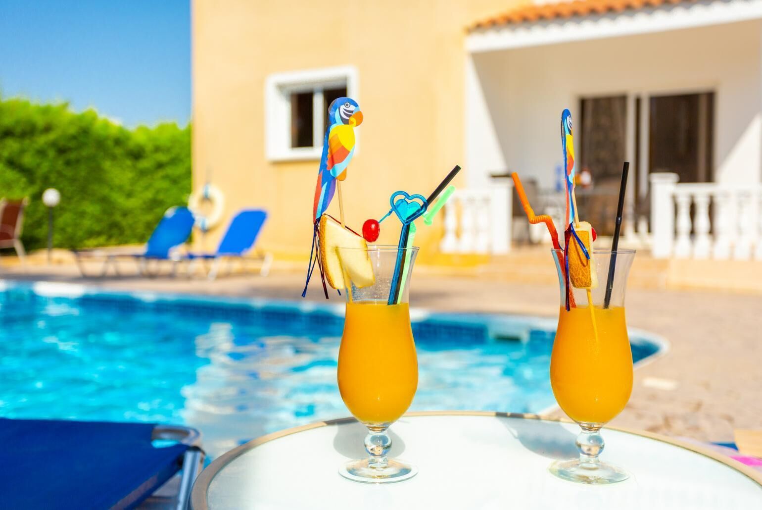 Dos cócteles con pajitas de colores y una decoración de loro, sobre una mesa junto a una piscina.