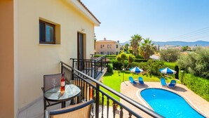 A balcony with a glass table and chairs, a pool with lounge chairs, and a view of a residential area with palm trees and houses. - Villa Halima Fostira (Poli Crysochous)