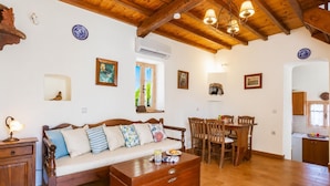Una habitación acogedora con techo de madera, un sofá con cojines, un aparador de madera, una mesa de comedor con sillas y una zona de cocina.