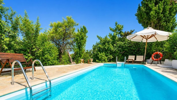 Una piscina azul clara con una escalera metálica, sillas de descanso de madera y una sombrilla blanca.