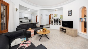 A modern living room with a black sofa, a coffee table, and a TV. There is a kitchen in the background with white cabinets and a dining area with a bar.