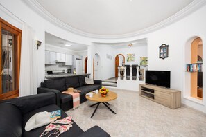 A modern living room with a black sofa, a coffee table, and a TV. There is a kitchen in the background with white cabinets and a dining area with a bar.
