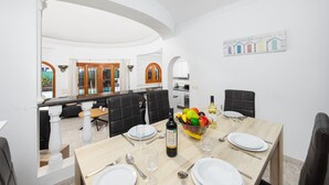 A dining area with a large table set for six, a fruit bowl, and a bottle of wine. There is a kitchen in the background with a view of the pool.