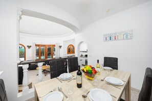 A dining area with a large table set for six, a fruit bowl, and a bottle of wine. There is a kitchen in the background with a view of the pool.