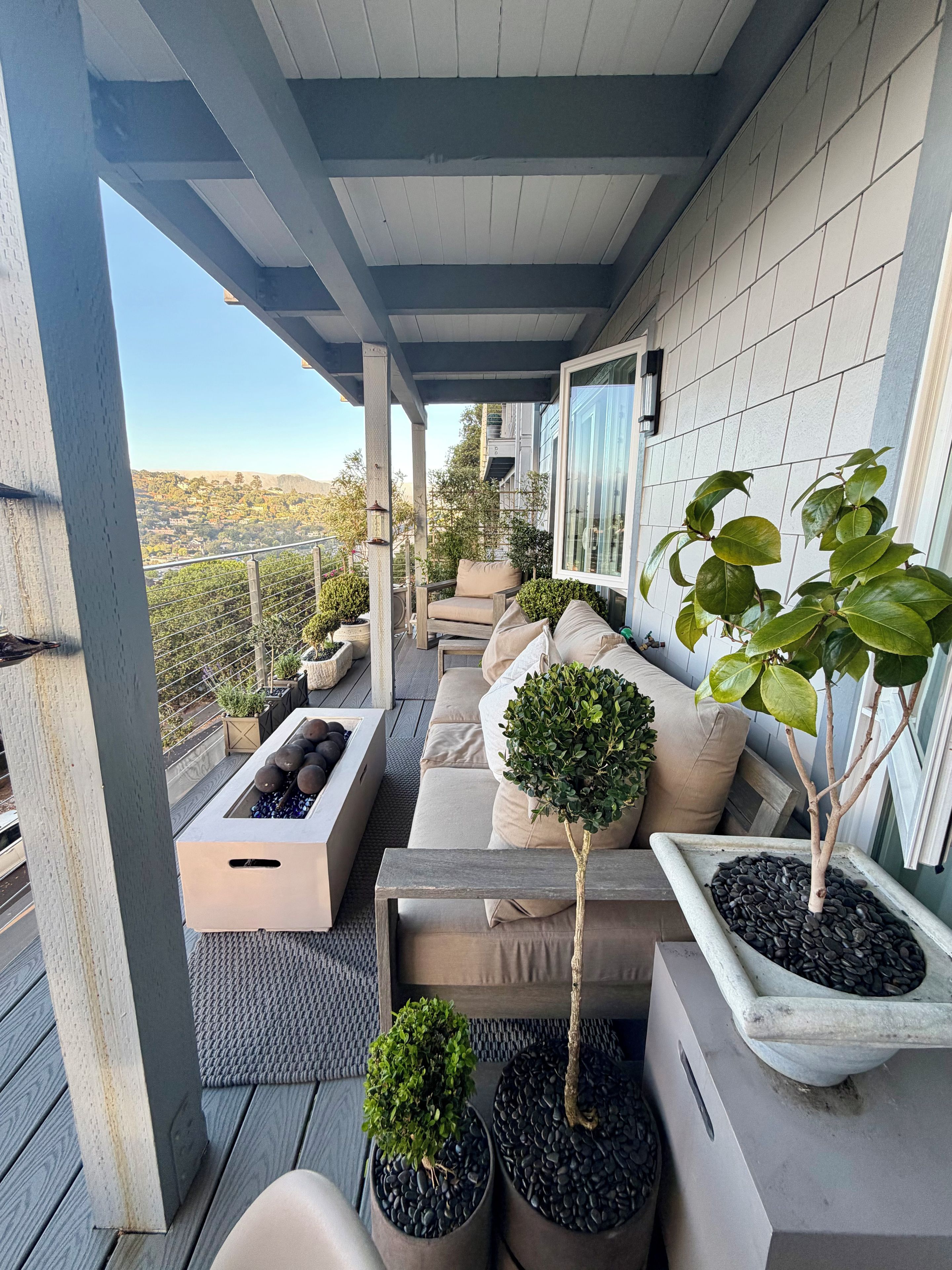 A covered outdoor seating area with a view of the cityscape, featuring a sofa, chairs, and potted plants.