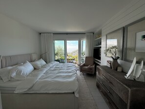 A modern bedroom with a large bed, a television, a dresser, and a view of the city.