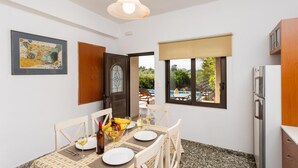A dining area with a table set for two, a bottle of wine, and a bowl of fruit. There is a wooden cabinet to the right and a framed picture on the wall. - Villa Maro Large Private Pool, A/C, WiFi (Kefalas)