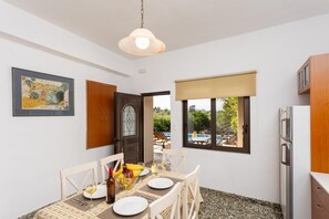 A dining area with a table set for two, a bottle of wine, and a bowl of fruit. There is a wooden cabinet to the right and a framed picture on the wall.