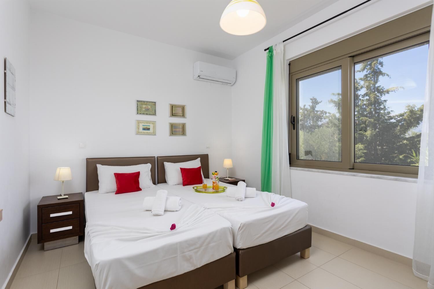 A double bed with white and red bedding, two bedside tables with lamps, a window with a view of trees, and an air conditioning unit on the wall.