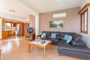 Un salon spacieux avec un canapé gris foncé, une table basse en bois et un téléviseur posé sur un meuble en bois. Un tableau est accroché au mur, et un appareil de climatisation est fixé au-dessus du canapé.