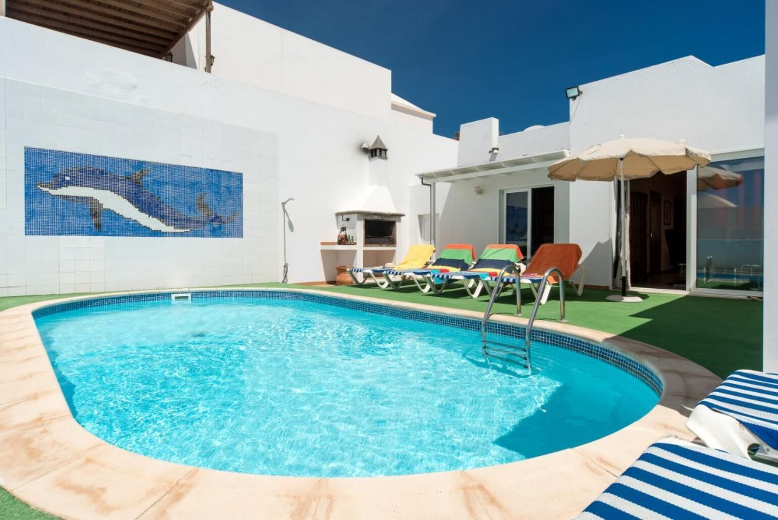 Une piscine entourée de chaises longues et agrémentée d’un parasol, le tout bordé d’un bâtiment blanc dont le mur est recouvert de carreaux et orné d’une mosaïque.