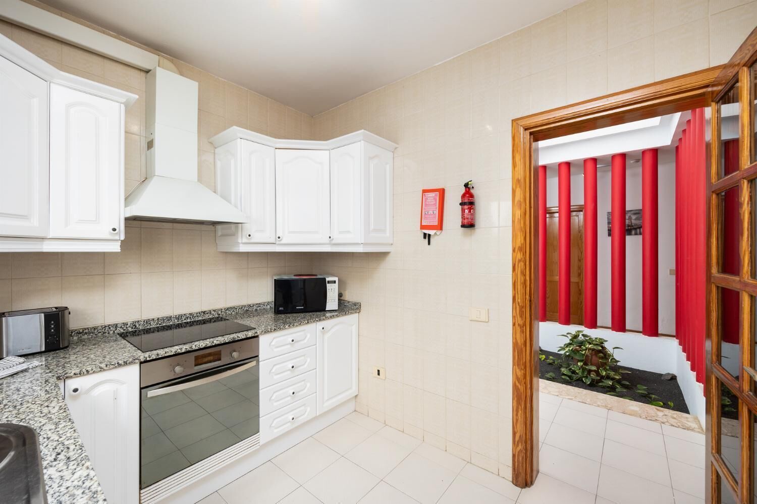 Una cocina con armarios blancos, un microondas, un tostador y una estufa. Hay un extintor en la pared y una puerta roja que da al exterior.