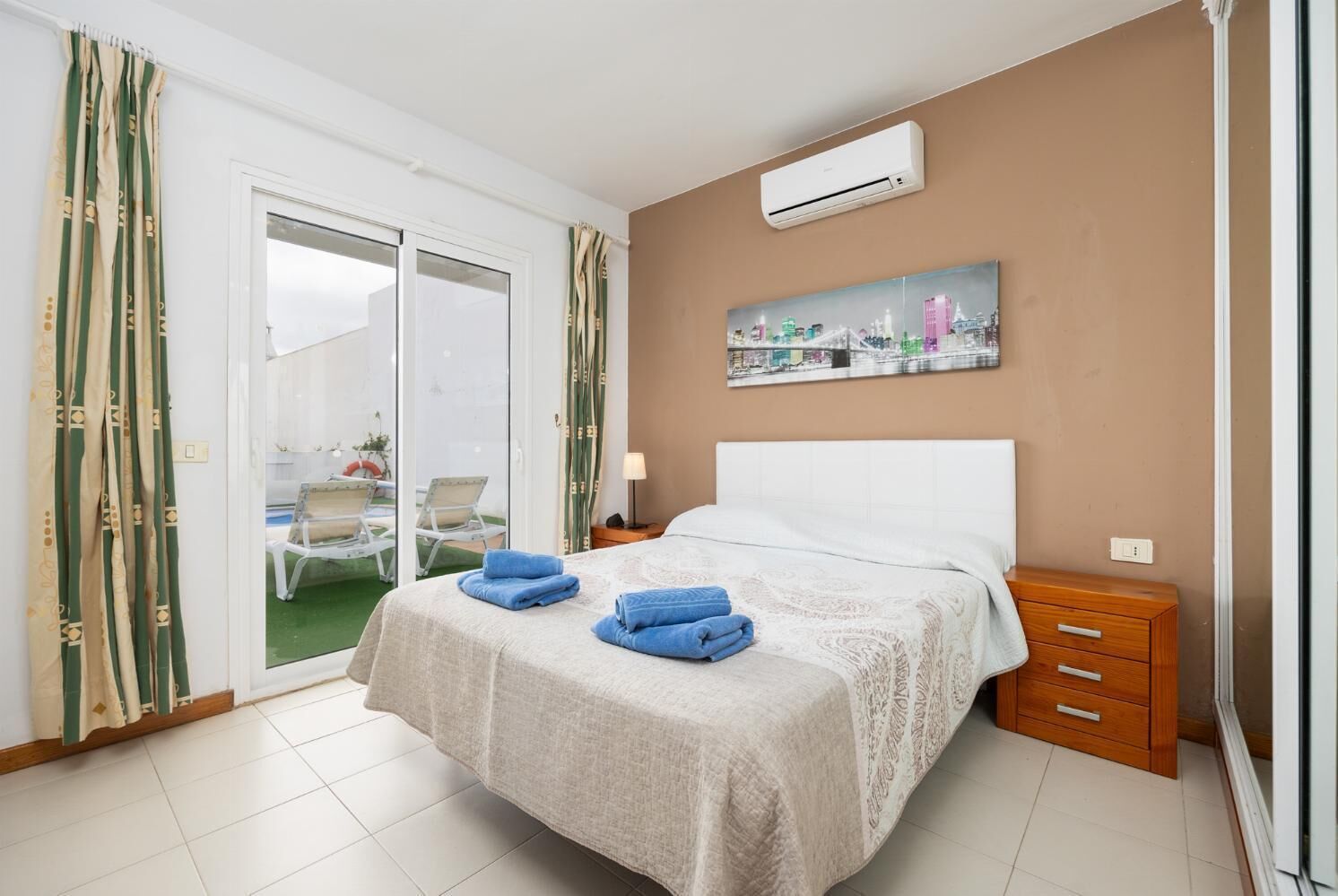 A hotel room with a bed, a nightstand, a wooden dresser, an air conditioning unit, and a cityscape painting on the wall.