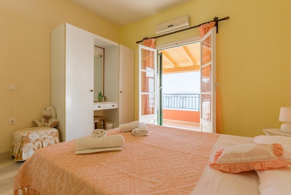 A bedroom with a bed, a dresser, a small table, a chair, a window with curtains, and an air conditioning unit.