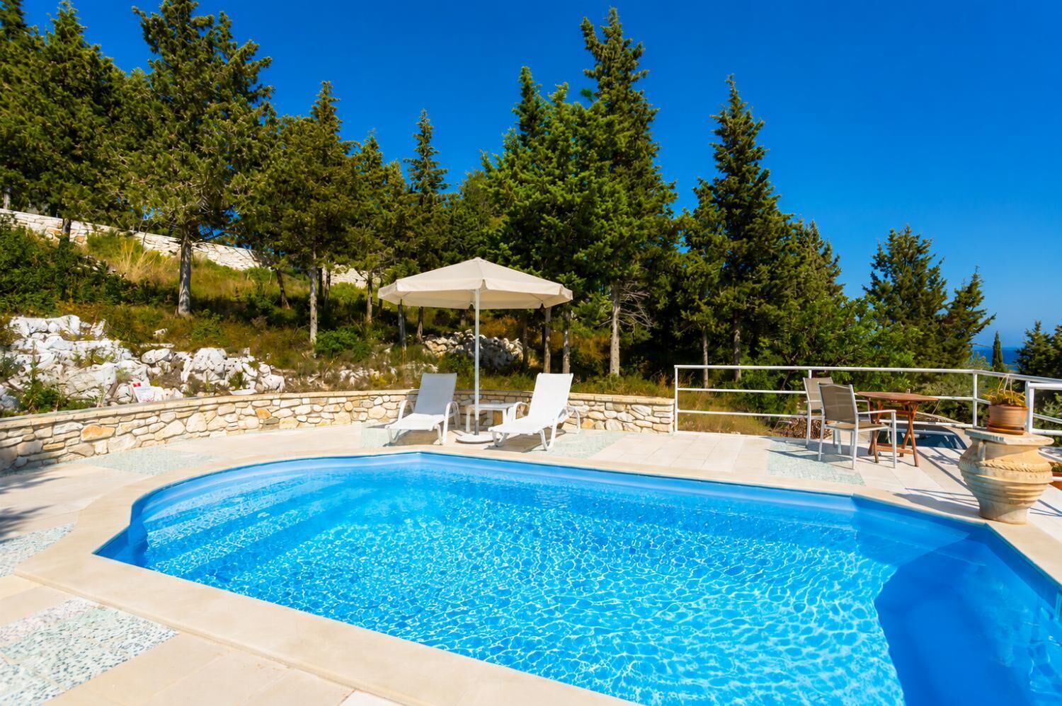 En swimmingpool med solstolar och ett bord, omgiven av träd och en stenmur.