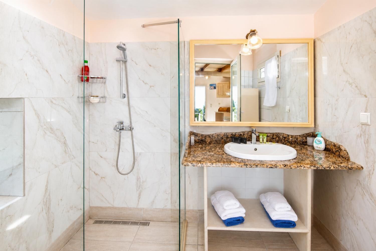 Une salle de bain dotée d’une douche à parois de verre, d’un mur recouvert de carreaux de marbre, d’un lavabo avec un plan de travail en granit et d’un miroir avec un luminaire intégré.