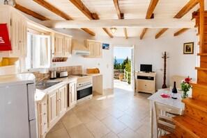 A kitchen with wooden beams, a dining area with a table and chairs, and a view of the sea through an open door. - Villa Windmill - One Bedroom Villa, Sleeps 2 (Paxi)