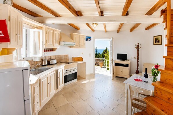 A kitchen with wooden beams, a dining area with a table and chairs, and a view of the sea through an open door.