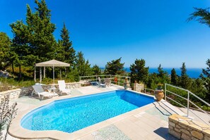 Piscina con sillones de descanso y pérgola, rodeada de árboles y con vista a un cuerpo de agua.