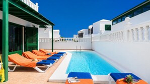Una piscina en la azotea con sillones de descanso, cielo azul despejado y edificios blancos.