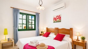 Una cama doble con ropa de cama roja y blanca, un cabecero de madera, dos mesitas de noche con lámparas, un ventanal con contraventanas verdes y un aire acondicionado instalado en la pared.