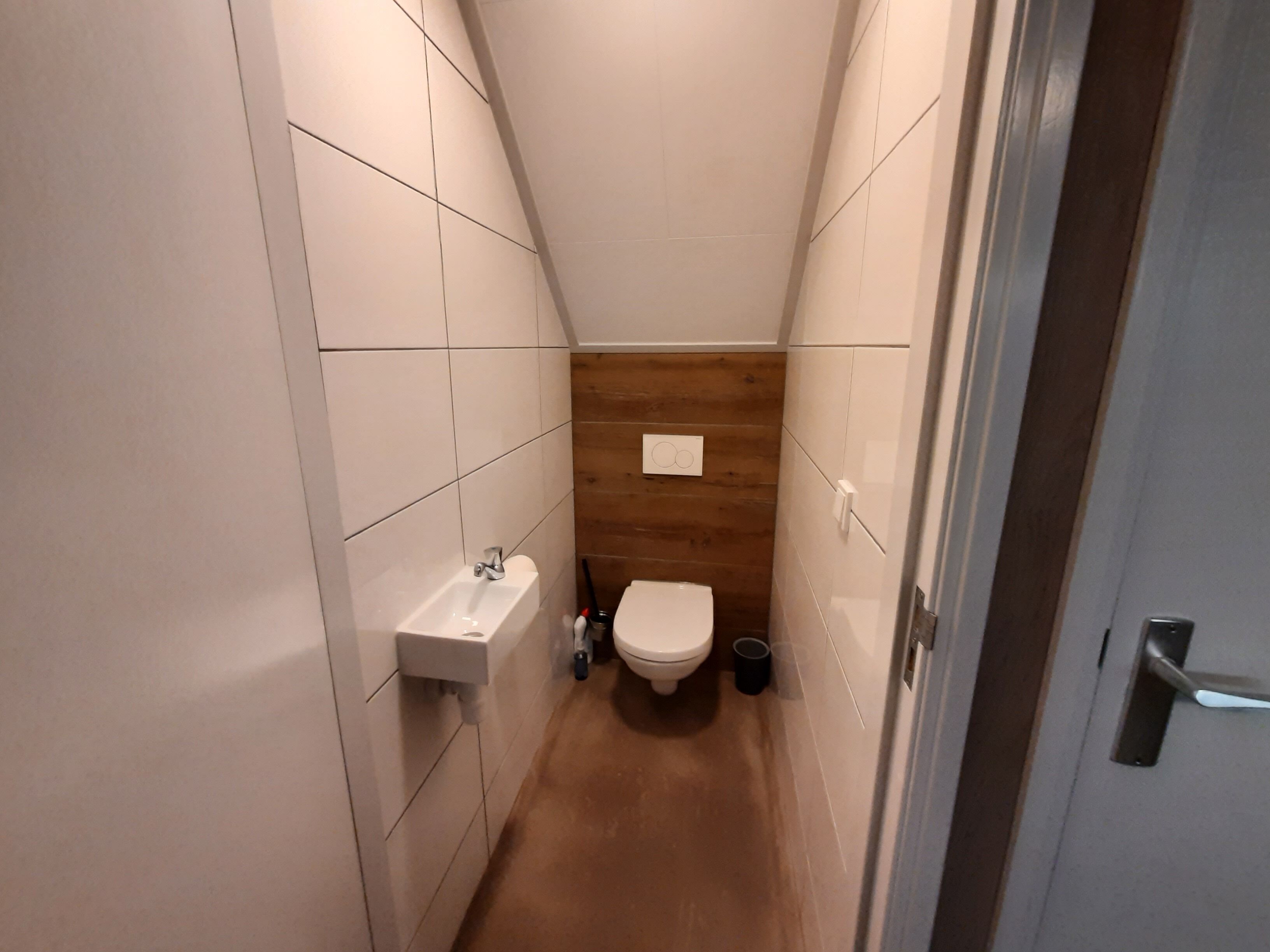 Une petite salle de bain aux murs recouverts de carreaux blancs, avec un mur d’accent en bois, un lavabo blanc et des toilettes.