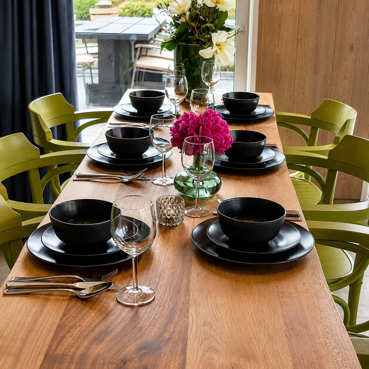 A dining table set with black bowls, wine glasses, and cutlery, surrounded by green chairs.