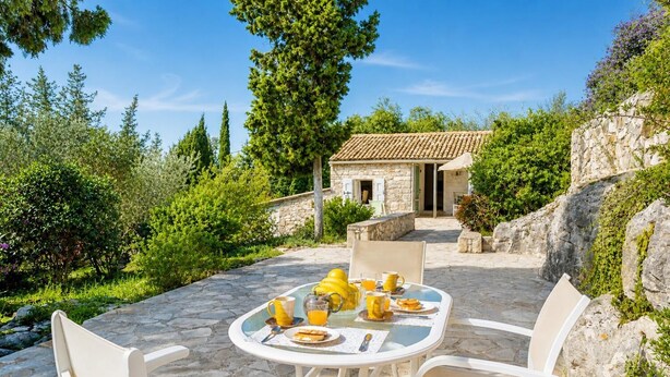 Un patio de piedra con una mesa dispuesta para el desayuno, rodeado de follaje y un edificio de piedra al fondo.