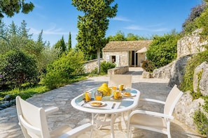 A stone patio with a table set for breakfast, surrounded by greenery and a stone building in the background. - Millers Cottage Large Private Pool, A/C, WiFi (Paxi)