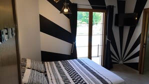 Ein Schlafzimmer mit einem Bett, schwarz-weiß gestreiften Kissen, einer gemusterten Tagesdecke, einer Deckenleuchte und Blick ins Freie durch ein Fenster.