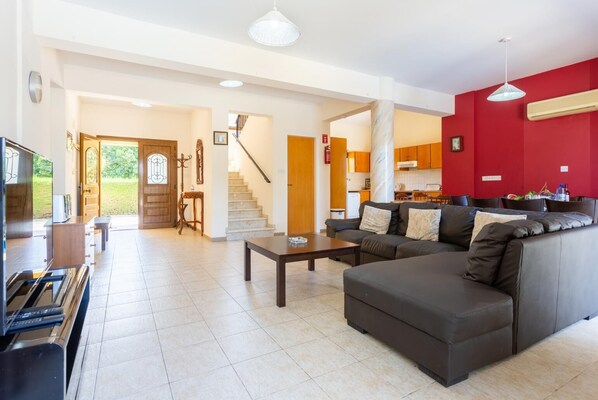 A spacious living area with a sofa, a coffee table, and a television. There is a staircase leading to a kitchen area with red walls and a dining table. - Villa Halima Stefanos (Poli Crysochous)