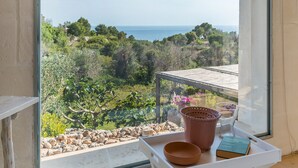 Un balcón con vistas al mar, una silla blanca, una bandeja con un tazón, un libro y una planta en maceta.