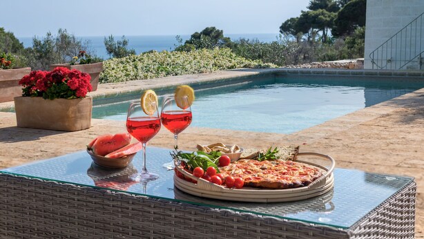 Mesa junto a la piscina con pizza, sandía y bebidas, con vista a la piscina y al océano.
