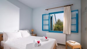A bedroom with a bed, a bedside table, a window with blue shutters, and a view of greenery outside. - Villa Eleni (Hersonissos)