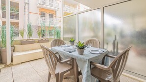 An outdoor dining area with a table set for four, surrounded by chairs, with potted plants and a view of a building with balconies.