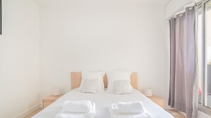 A neatly made bed with white linens and two towels, a wooden headboard, bedside lamps, and a window with curtains.
