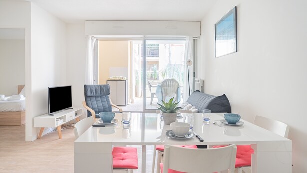 A modern living room with a white sofa, a dining table with chairs, a TV, and a balcony with a view.