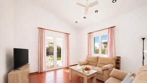 Un salón con televisor, sofá, mesa con fruta y un balcón con cortinas.