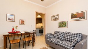 A living room with a sofa, dining table, chairs, and kitchen in the background. - Foteini Apartment - Two Bedroom Apartment, Sleeps 2 (Corfu)