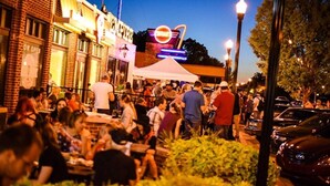 A crowded outdoor dining area with brick buildings and a neon sign in the background.