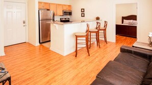 A modern kitchen with a central island, wooden cabinets, and stainless steel appliances. There is a seating area with a dark leather couch and a small table.