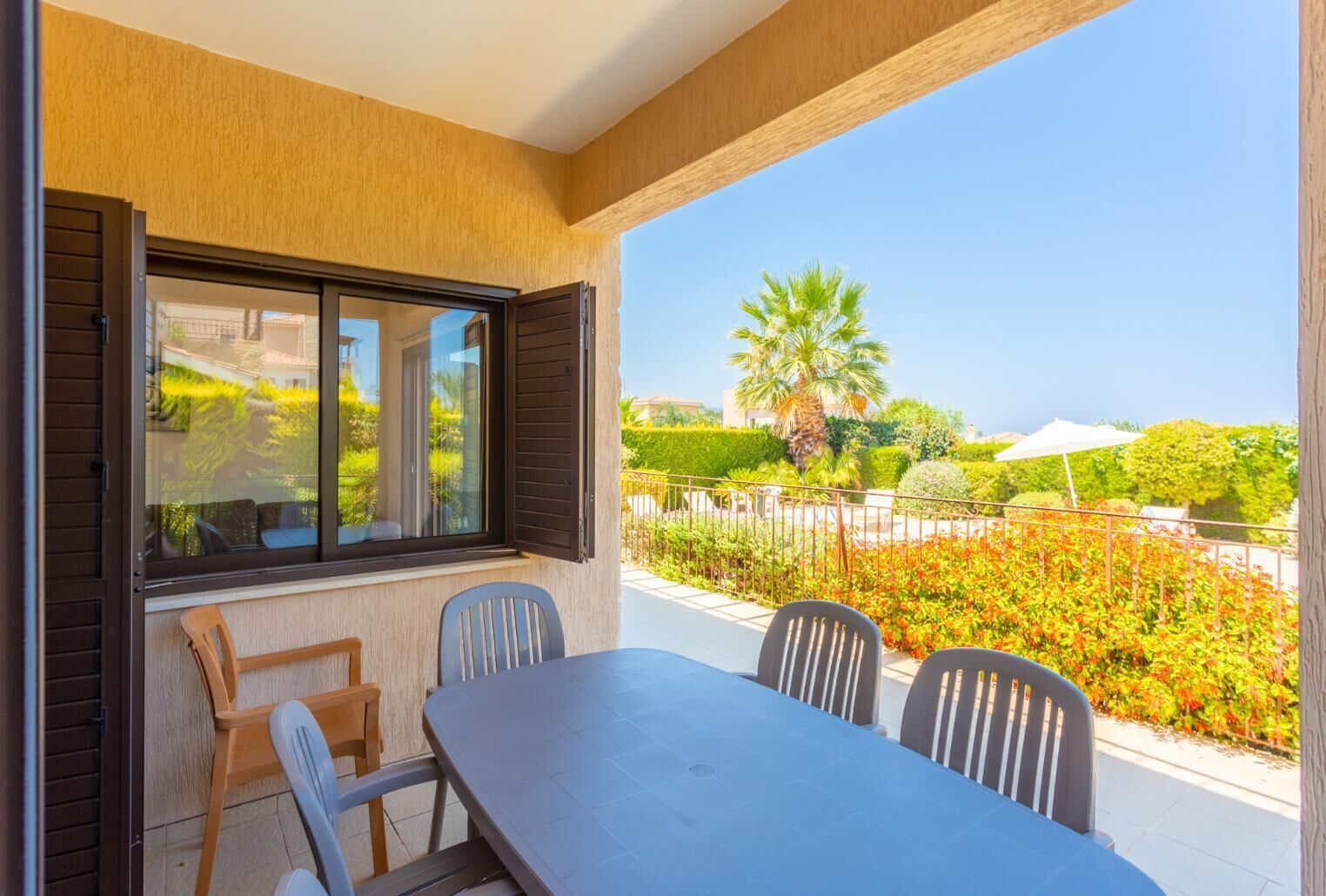 A balcony with a table and chairs, a view of a garden with palm trees and shrubs, and a clear blue sky.