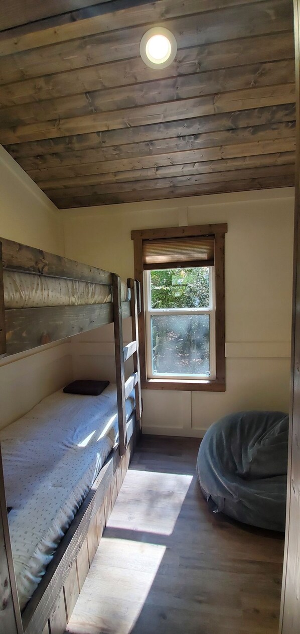 Ein kleiner, gemütlicher Raum mit einem Etagenbett, einem Einzelbett, einem Fenster mit Blick auf Grünflächen und einer Deckenleuchte.