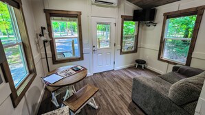 Una habitación con techo de madera, un sofá, un banco, una mesa con camino de mesa y ventanas amplias con vistas a zonas verdes.
