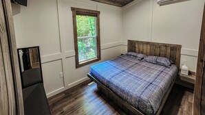 Un dormitorio con techo de madera, ventana con vista a la vegetación, cama con colcha estampada y unidad de aire acondicionado blanca.
