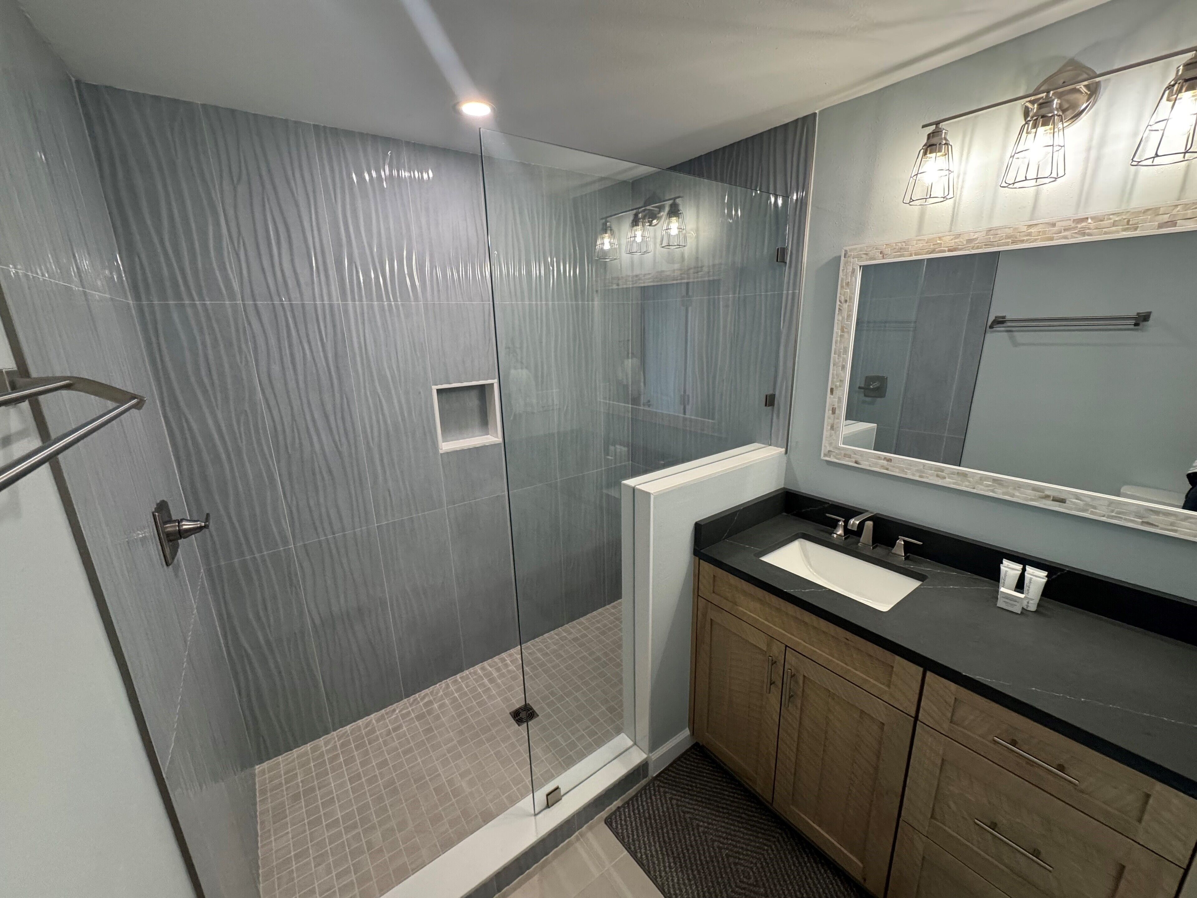 A modern bathroom with a glass shower enclosure, a large mirror, and a dark countertop.
