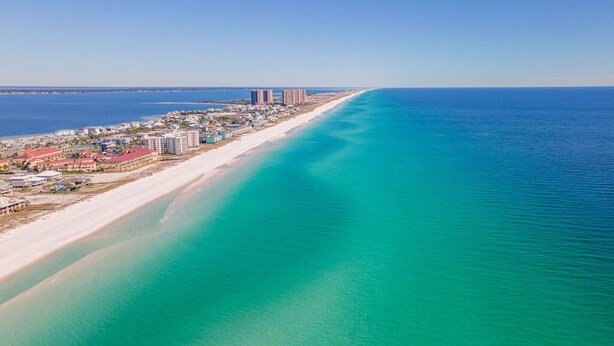 An aerial view of a coastal area with a long sandy beach, a clear blue ocean, and a row of buildings along the shore.
