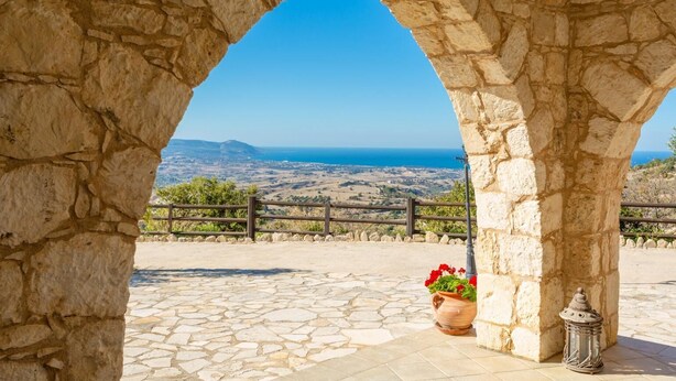 Un arco de piedra que conduce a una terraza con vistas al mar y colinas al fondo.